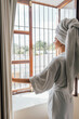 © Berena Alvarez/Stocksy - Young woman in white bathrobe take air at the window