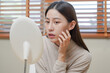 © KMPZZZ - Dermatology, puberty asian young woman, girl looking into mirror, allergy presenting an allergic reaction from cosmetic, red spot or  rash on face. Beauty care from skin problem by medical treatment.