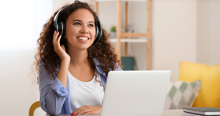 Wall Mural - African-American woman with headphones listening to music while working on laptop at home