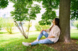 Young Asian Thai, Vietnamese or Chinese woman in oversize shirt, jeans and glasses sitting relaxing recreating in shadow under green tree on weekend or holiday in city park on summer sunny day. 