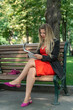 © somemeans - Beautiful young woman in red dress, reading newspaper on bench in park. Attractive girl in park.