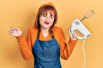 Poster - Redhead young woman holding pastry blender electric mixer screaming proud, celebrating victory and success very excited with raised arm