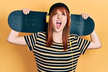 Wall Mural - Redhead young woman holding skate wearing headphones angry and mad screaming frustrated and furious, shouting with anger looking up.