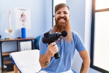 Poster - Young redhead man wearing physiotherapist uniform holding percussion massage pistol at physiotherapy clinic