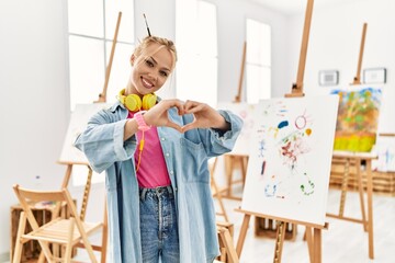 Poster - Young caucasian girl at art studio smiling in love doing heart symbol shape with hands. romantic concept.