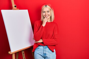 Sticker - Beautiful blonde woman standing by painter easel stand smiling looking confident at the camera with crossed arms and hand on chin. thinking positive.