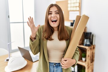 Canvas Print - Young brunette woman holding paper blueprints at the office celebrating victory with happy smile and winner expression with raised hands