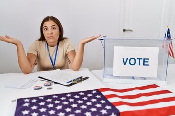 Canvas Print - Young brunette woman at political election sitting by ballot clueless and confused expression with arms and hands raised. doubt concept.