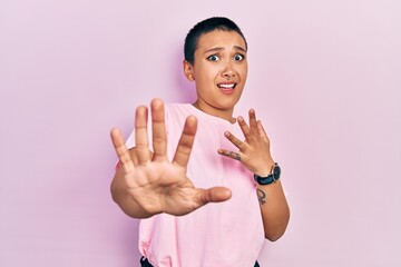 Sticker - Beautiful hispanic woman with short hair wearing casual pink t shirt afraid and terrified with fear expression stop gesture with hands, shouting in shock. panic concept.