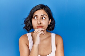 Poster - Young hispanic woman standing over blue background looking confident at the camera with smile with crossed arms and hand raised on chin. thinking positive.