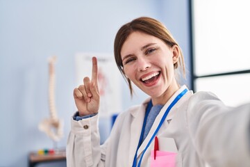 Sticker - Young physiotherapist woman working at pain recovery clinic smiling with an idea or question pointing finger with happy face, number one