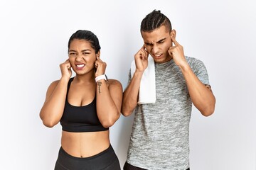 Young latin couple wearing sportswear standing over isolated background covering ears with fingers with annoyed expression for the noise of loud music. deaf concept.