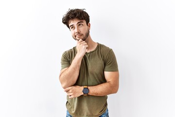 Canvas Print - Hispanic man standing over isolated white background looking confident at the camera smiling with crossed arms and hand raised on chin. thinking positive.