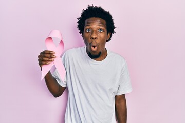 Poster - Young african american man holding pink cancer ribbon scared and amazed with open mouth for surprise, disbelief face