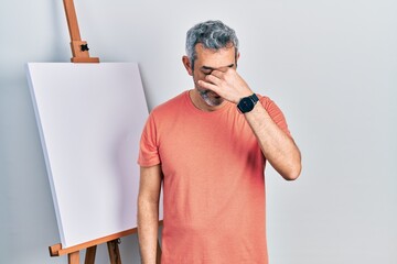 Canvas Print - Handsome middle age man with grey hair standing by painter easel stand tired rubbing nose and eyes feeling fatigue and headache. stress and frustration concept.