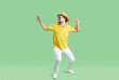 Happy young man in summer clothes, hat and sunglasses dance on green studio background. Overjoyed male in casualwear have fun make funny dancer moves. Entrainment and party.