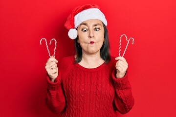 Poster - Middle age hispanic woman wearing christmas hat holding candy making fish face with mouth and squinting eyes, crazy and comical.