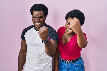 Poster - Young african american couple standing over pink background angry and mad raising fist frustrated and furious while shouting with anger. rage and aggressive concept.