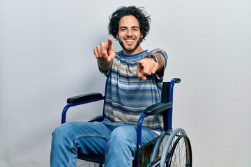 Poster - Handsome hispanic man sitting on wheelchair pointing to you and the camera with fingers, smiling positive and cheerful