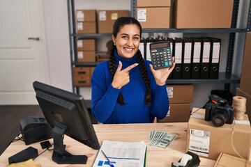 Poster - Young hispanic woman working at small business ecommerce holding calculator smiling happy pointing with hand and finger