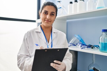 Wall Mural - Young hispanic woman wearing scientist uniform writing on clipboard at laboratory