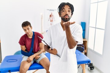 Wall Mural - Young hispanic man working at pain recovery clinic with a man with broken arm looking at the camera blowing a kiss with hand on air being lovely and sexy. love expression.