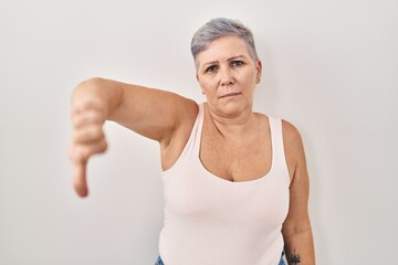 Poster - Middle age caucasian woman standing over white background looking unhappy and angry showing rejection and negative with thumbs down gesture. bad expression.