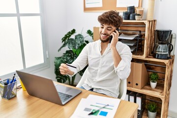 Wall Mural - Young arab man talking on the smartphone and holding credit card working at office