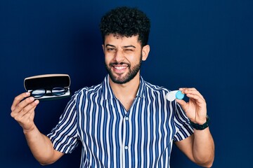 Canvas Print - Young arab man with beard holding glasses and contact lenses winking looking at the camera with sexy expression, cheerful and happy face.