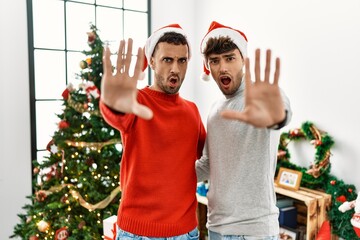 Sticker - Young gay couple standing by christmas tree wearing hat doing stop gesture with hands palms, angry and frustration expression