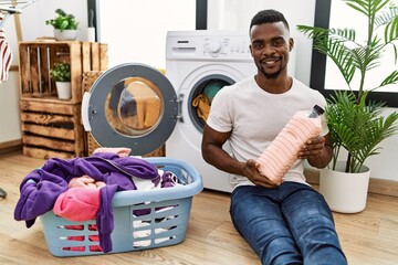 Wall Mural - Young african man holding detergent bottle at laundry room looking positive and happy standing and smiling with a confident smile showing teeth