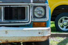 The Front Of A Blue Car Including The Grate, Headlight And A Rusted Bumper With A Portion Of A Yellow Car In The Background.