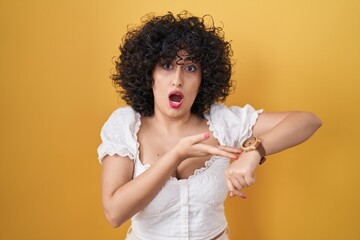 Sticker - Young brunette woman with curly hair standing over yellow background in hurry pointing to watch time, impatience, upset and angry for deadline delay