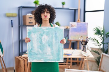 Canvas Print - Young brunette woman with curly hair holding painter canvas puffing cheeks with funny face. mouth inflated with air, catching air.