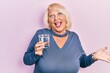 Middle age blonde woman drinking glass of water celebrating achievement with happy smile and winner expression with raised hand