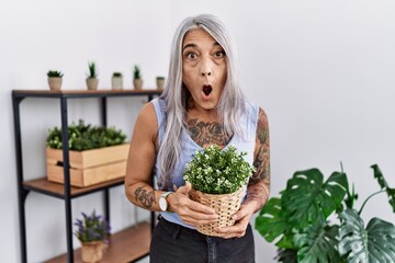 Poster - Middle age grey-haired woman holding green plant pot at home in shock face, looking skeptical and sarcastic, surprised with open mouth