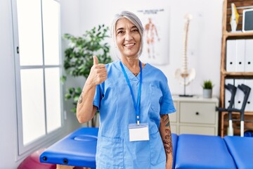 Poster - Middle age grey-haired woman wearing physiotherapist uniform at medical clinic doing happy thumbs up gesture with hand. approving expression looking at the camera showing success.