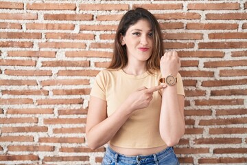 Wall Mural - Young brunette woman standing over bricks wall in hurry pointing to watch time, impatience, looking at the camera with relaxed expression