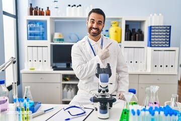 Canvas Print - Young hispanic man with beard working at scientist laboratory cheerful with a smile of face pointing with hand and finger up to the side with happy and natural expression on face