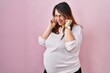 Pregnant woman standing over pink background covering ears with fingers with annoyed expression for the noise of loud music. deaf concept.