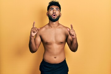 Canvas Print - Arab man with beard wearing swimwear shirtless amazed and surprised looking up and pointing with fingers and raised arms.