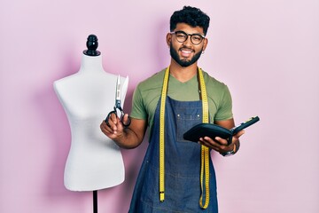 Canvas Print - Arab man with beard dressmaker designer holding scissors and sewing kit smiling with a happy and cool smile on face. showing teeth.