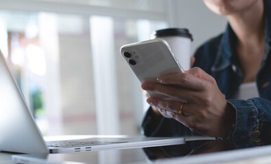 Poster - Young smart asian business woman sitting at table, drinking coffee and using mobile phone with laptop computer on table, have a coffee break during working at home office