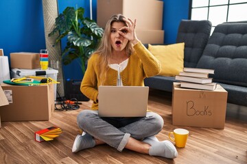 Poster - Young woman sitting on the floor at new home using laptop doing ok gesture shocked with surprised face, eye looking through fingers. unbelieving expression.