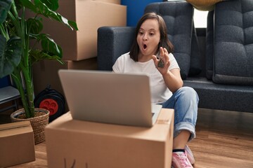 Wall Mural - Hispanic girl with down syndrome sitting on the floor at new home with laptop scared and amazed with open mouth for surprise, disbelief face