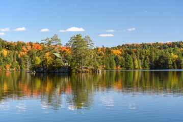 Wall Mural - Cabin perched on a small rocky island in the middle of a lake surrounded by forest on a sunny autumn day. People on a canoe are near the island. Stunning fall foliage. Algonquin Park, ON, Canada.