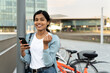© Maria Vitkovska - Attractive smiling Indian woman holding mobile phone, shopping online looking at camera outdoors. Beautiful happy asian tourist with backpack walking on city street, copy space. Travel concept