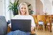 © baranq - Mid adult woman at home sitting on sofa using laptop working remotely