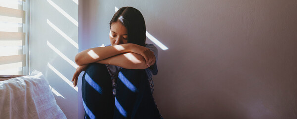 Asian women are sitting hugging their knees in bed. Feeling sad, disappointed in love In the dark bedroom and sunlight from the window through the blinds.