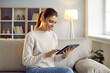 Smiling young Caucasian woman sit on sofa use modern tablet gadget. Happy millennial girl relax at home shopping online or browse internet on pad device. Technology and communication.